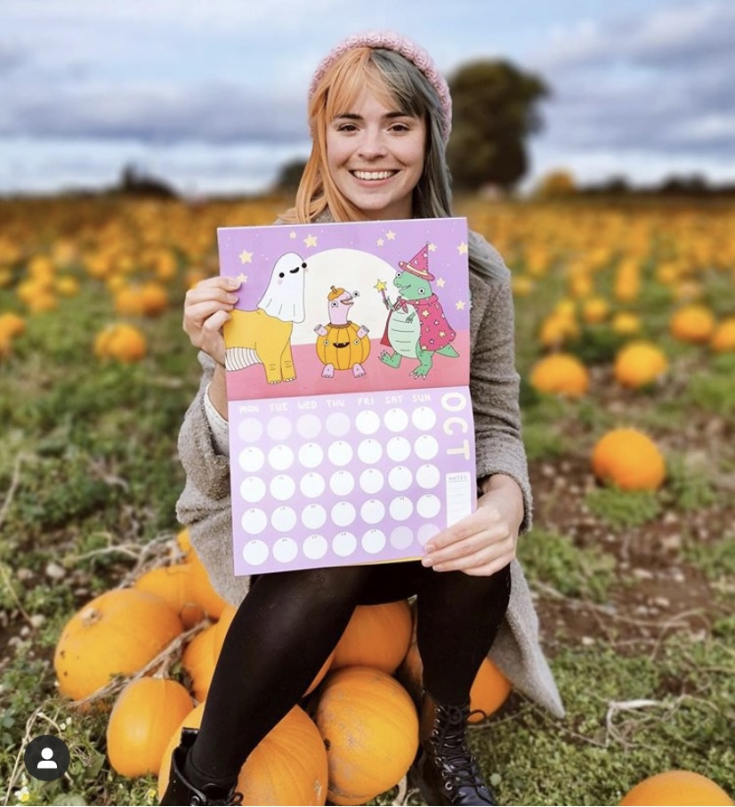 Girl sat on a pile of pumpkins in a pumpkin field holding a calendar opened to the October page. 