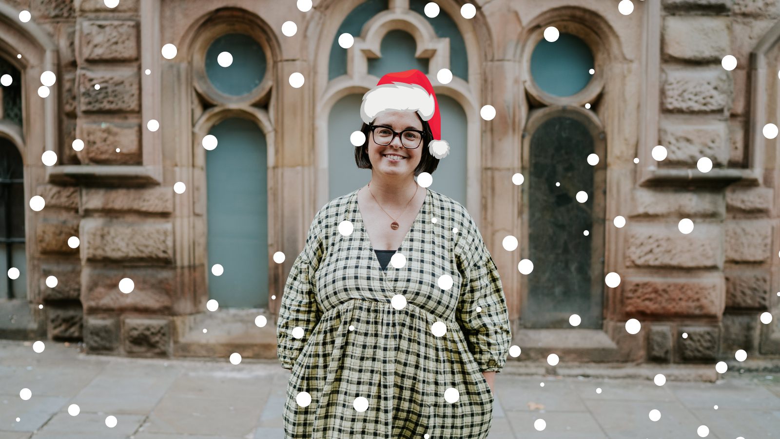 Smiling woman standing with her hands in her pockets standing in front of a building. There is a Santa hat on her head and an overlay of snow on the image.